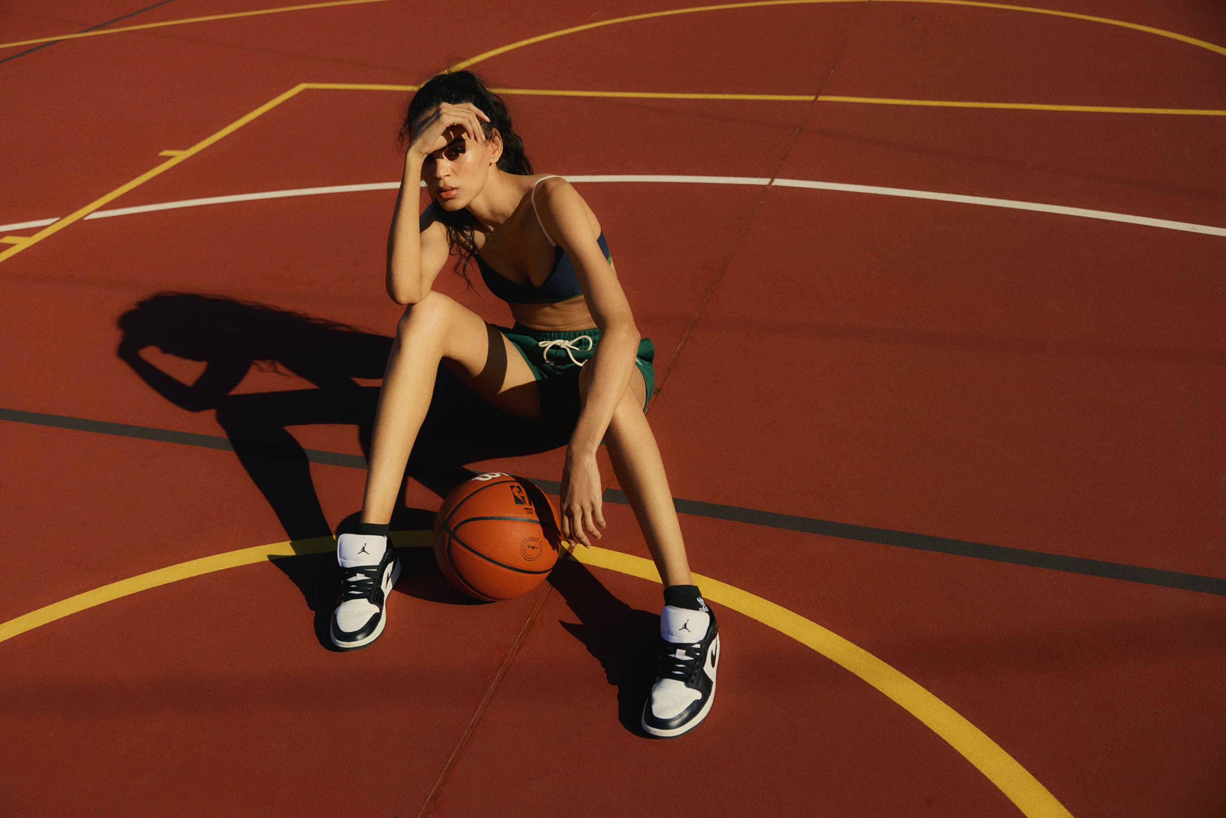Young woman in sports bra and shorts sits on a basketball court with a basketball, wearing athletic shoes in the sunlight.
