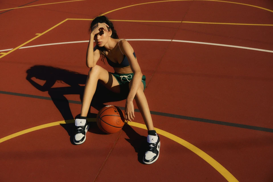 Young woman in sports bra and shorts sits on a basketball court with a basketball, wearing athletic shoes in the sunlight.