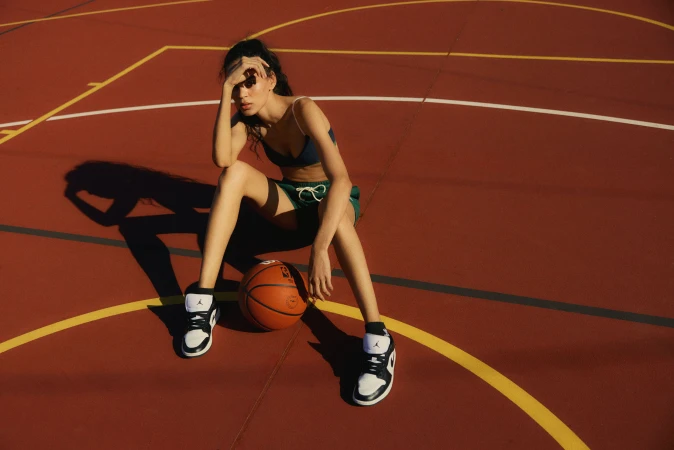 Young woman in sports bra and shorts sits on a basketball court with a basketball, wearing athletic shoes in the sunlight.