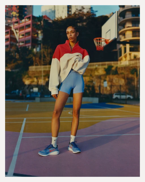 Woman stands confidently on colorful court wearing athletic apparel, including running shoes, against urban background with blue sky. 