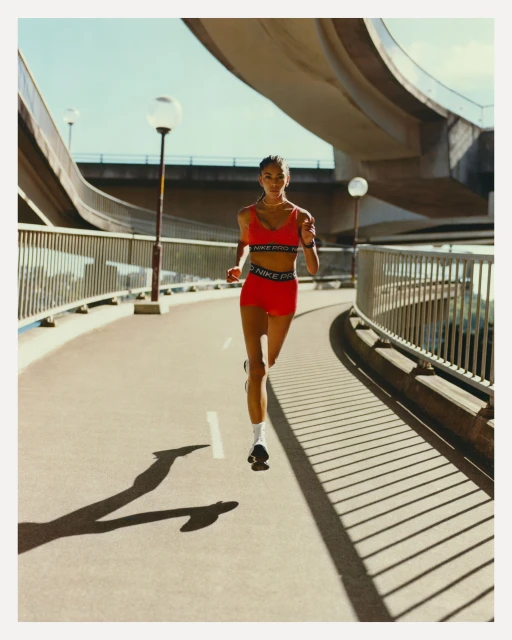 Athletic woman in red sportswear runs along a city walkway with overpass structures under a bright blue sky in an eye-level shot.