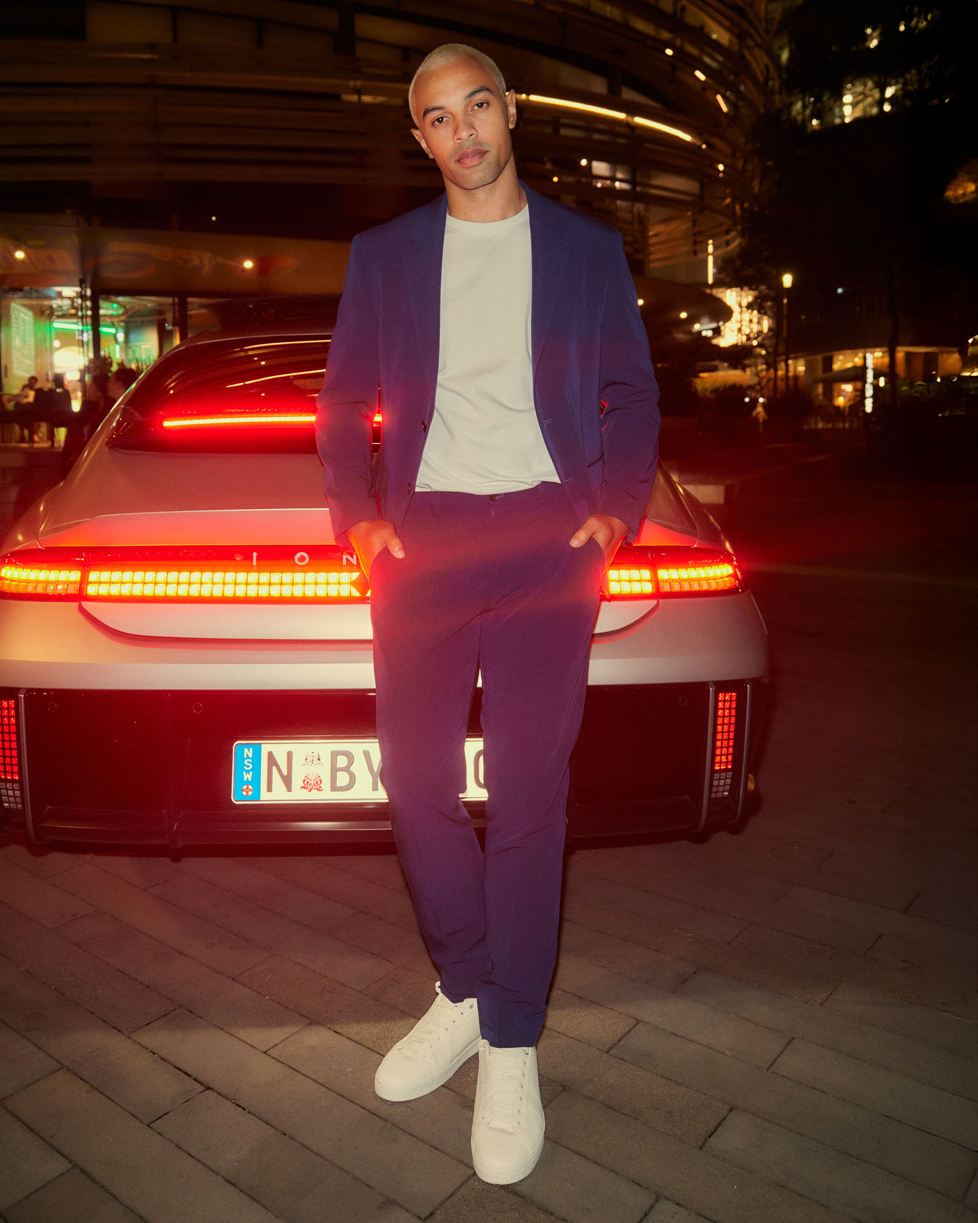 Man posing near a silver car in a modern urban setting at night, wearing a blue suit and white sneakers, illuminated by streetlights.