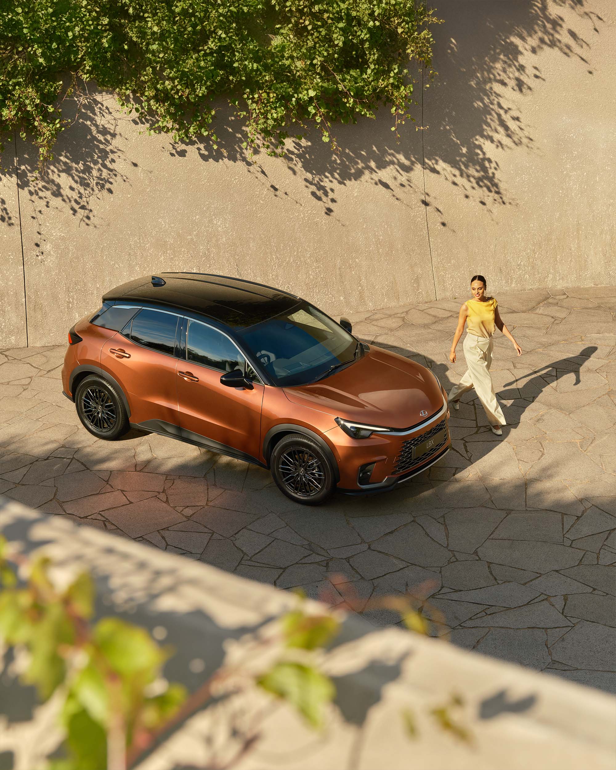 Bronze-colored SUV parked on a stone driveway, woman walking in background, sunny day with shadows on the wall and vehicle.