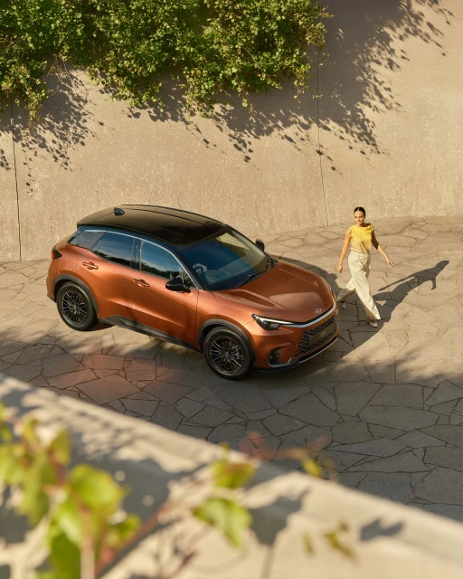 Bronze-colored SUV parked on a stone driveway, woman walking in background, sunny day with shadows on the wall and vehicle.