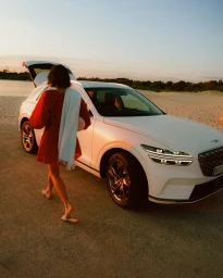 Woman in red dress walking toward white SUV with open trunk parked near water at sunset