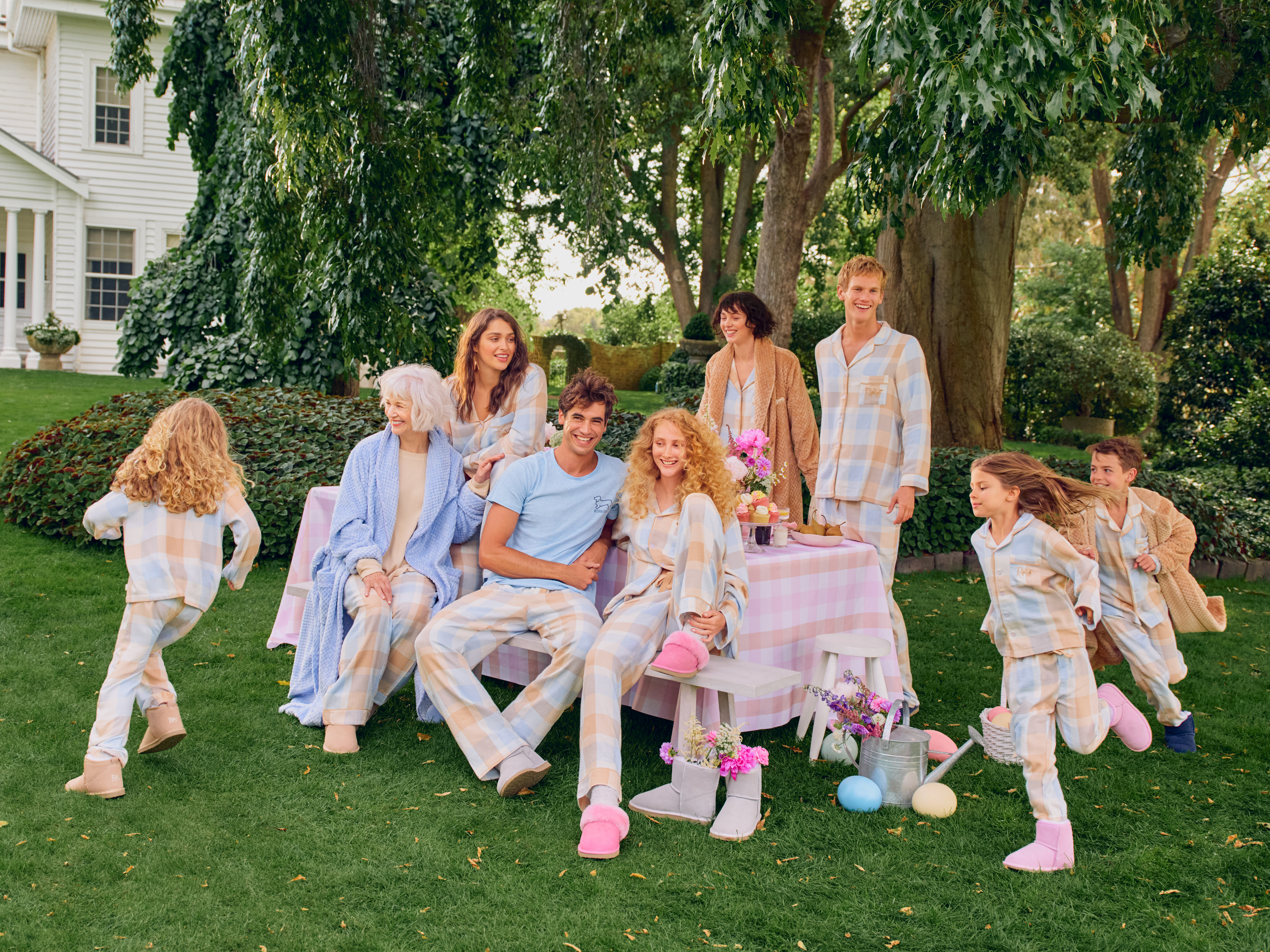 Family in matching pajamas gathers outdoors on a lawn for Easter with a pink tablecloth, flowers, easter eggs, and a white house.