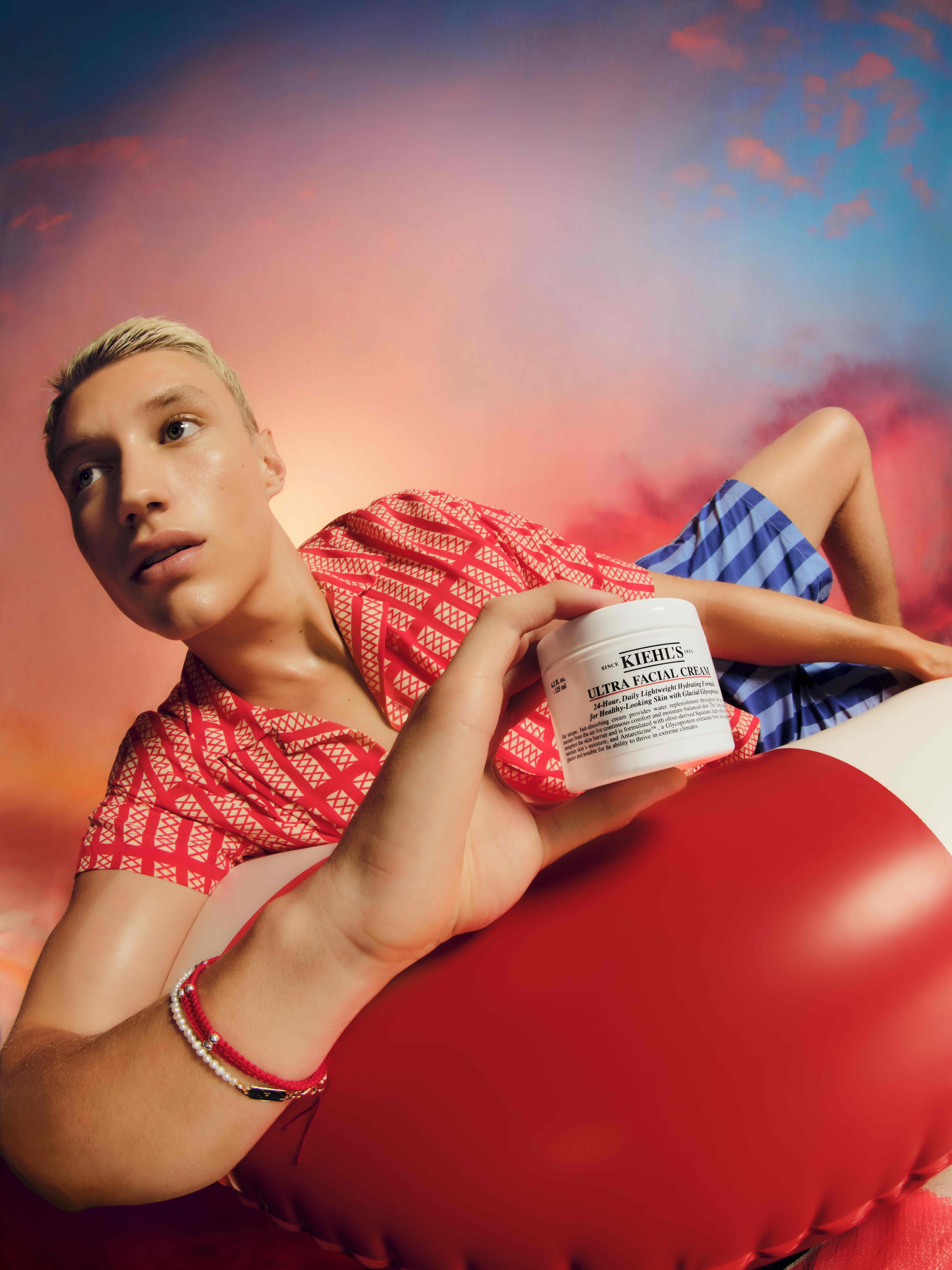 Person lies on red and white inflatable holding a white Kiehl's Ultra Facial Cream jar in a beauty product shot with a soft sky background.