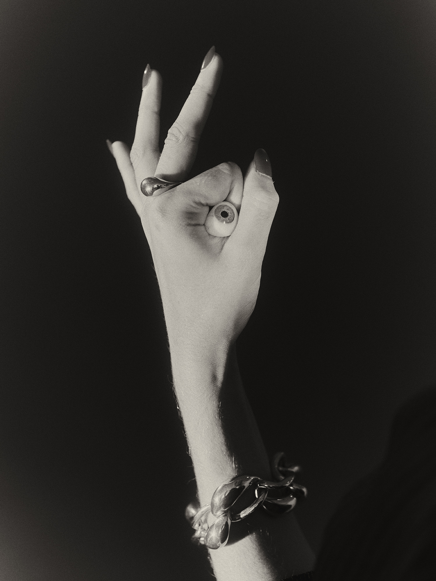 Monochrome studio shot of a female hand with painted nails making an OK sign, an eyeball in the circle, and a bracelet on the wrist.