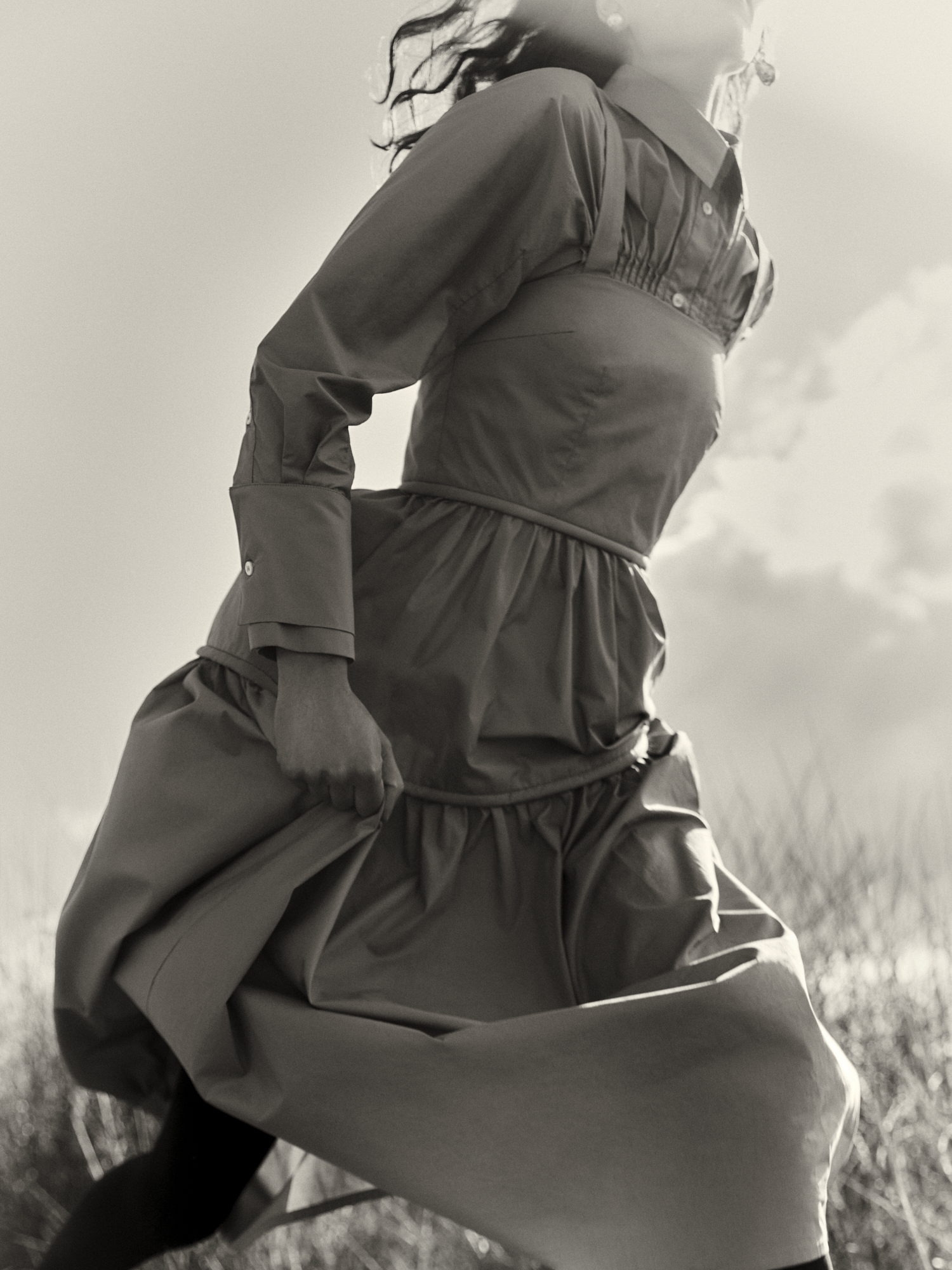 Monochromatic shot of a woman in motion, wearing a dress, with a neutral color palette and a background featuring grass and the sky.