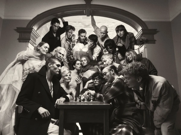 Monochromatic shot of a large group of people gathered around a chessboard, creating a sense of mystery and anticipation in an indoor setting.