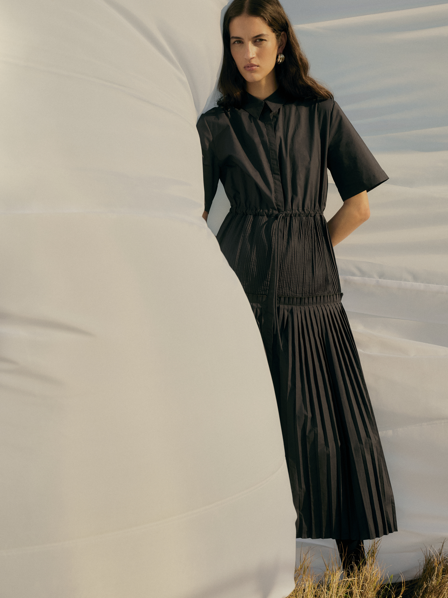 A woman models a black, pleated dress while standing in front of a white sheet with neutral sunlight and a grassy ground.