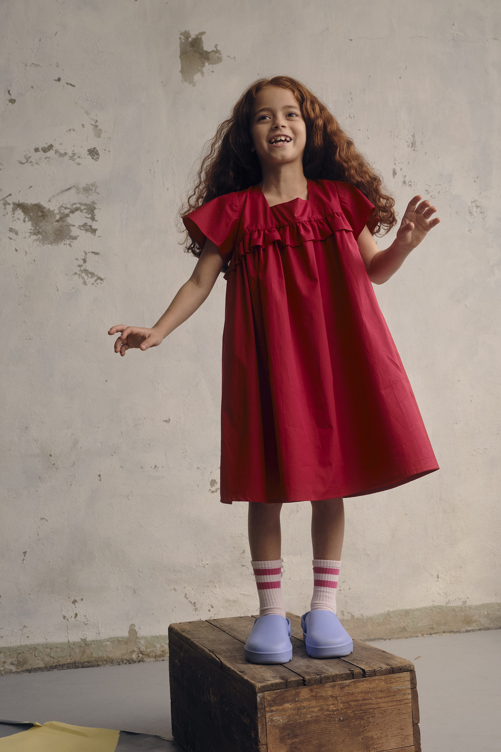 A young girl with curly red hair, wearing a red dress, stands on a wooden crate in a studio with a mottled background.