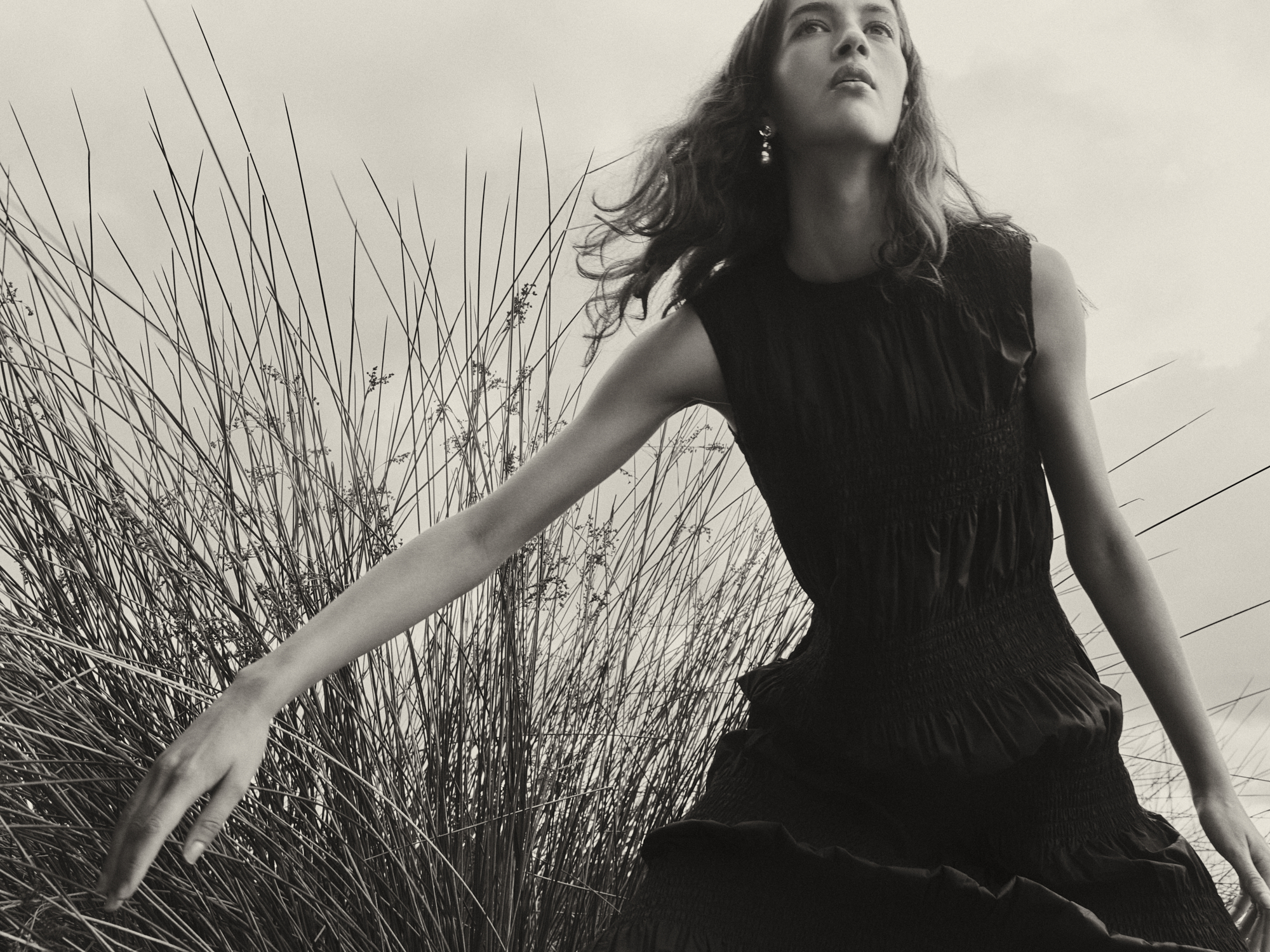 Monochrome fashion shot: a woman in a dress poses outdoors, tall grass in the background, creating a vintage and artistic visual.