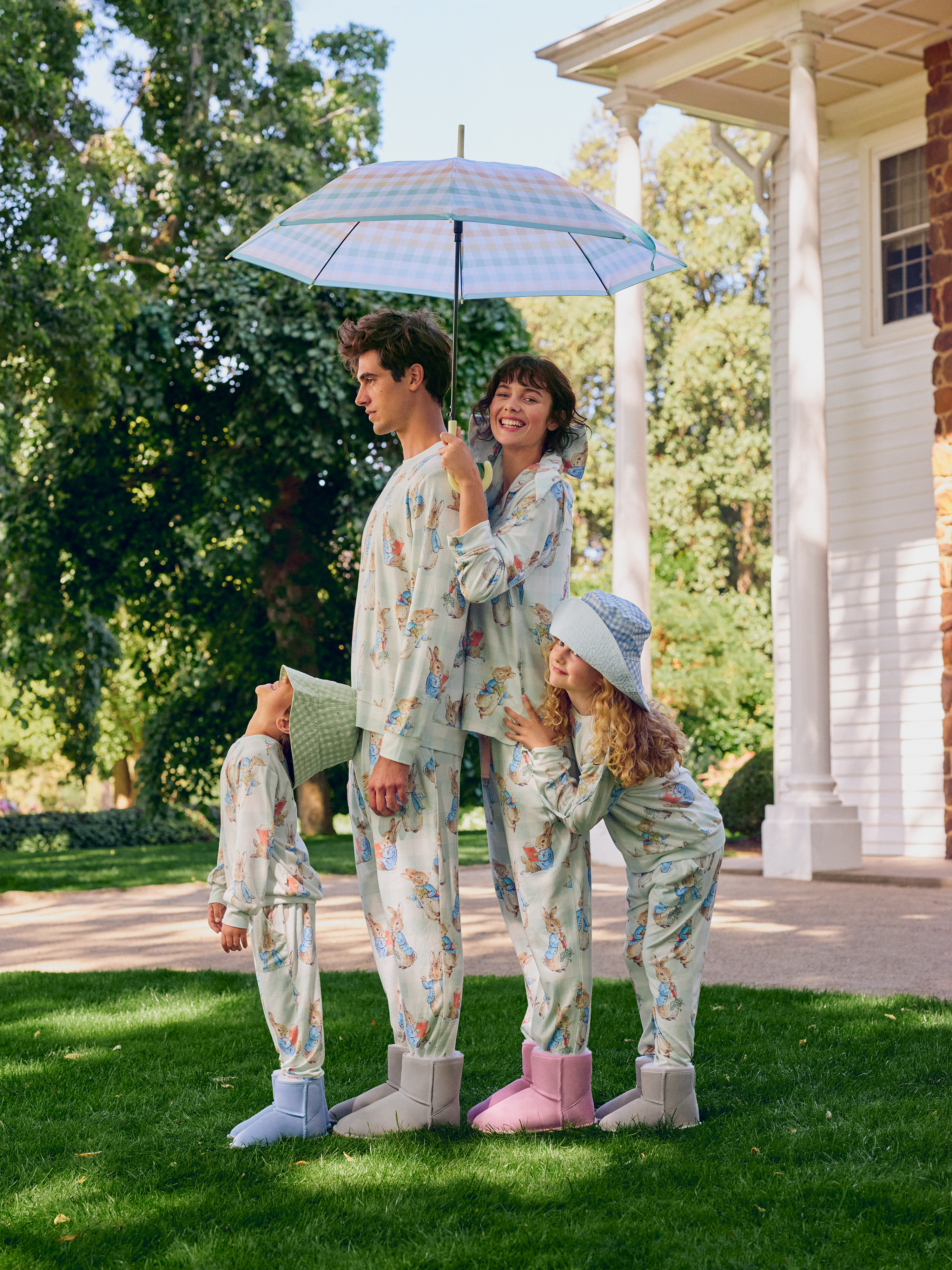 Family of four stands on a lawn under an umbrella, wearing matching pajamas and boots, with a large white house in the background.