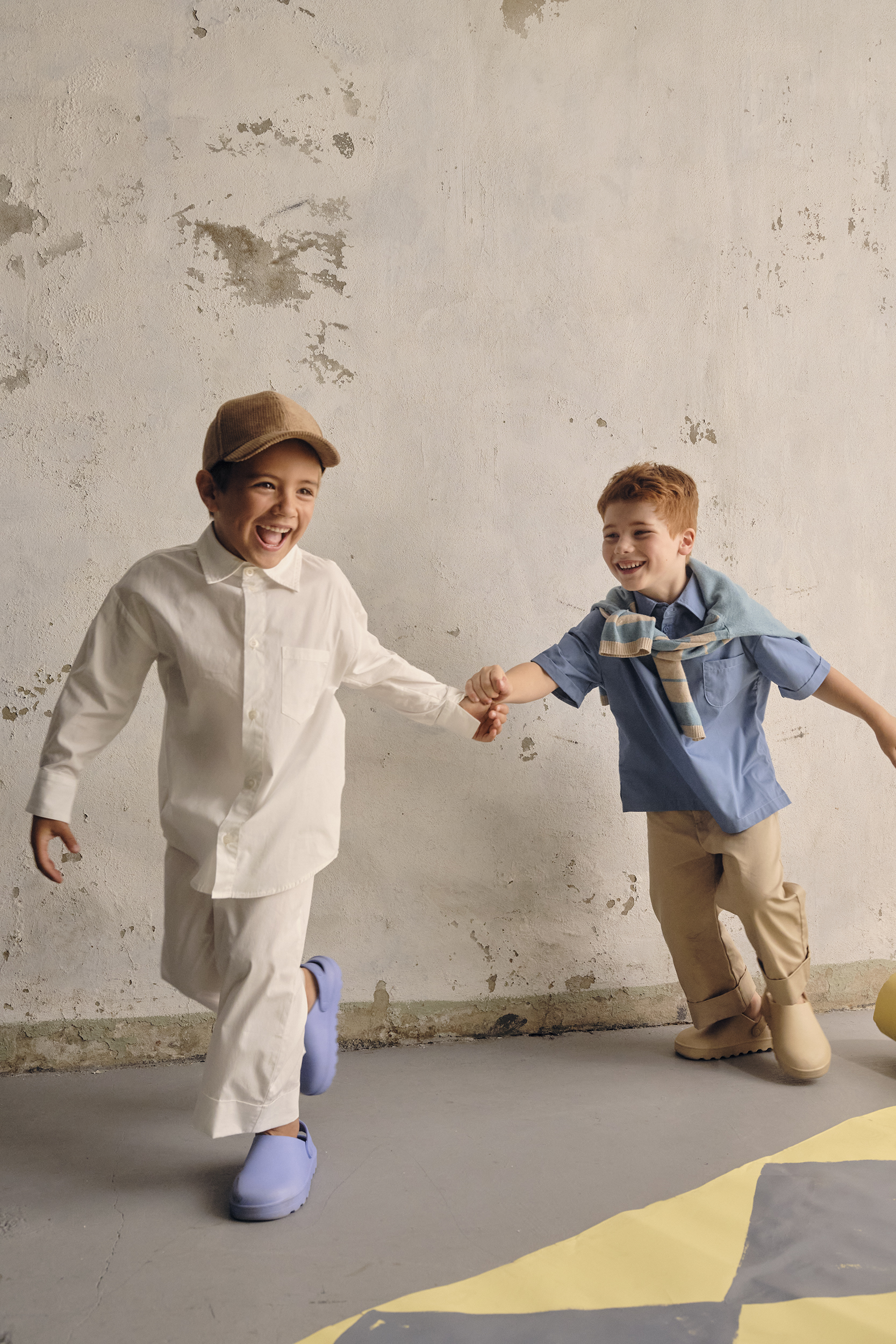 Two smiling children are running and holding hands in a room with a textured wall. The kids are wearing casual clothing and shoes.