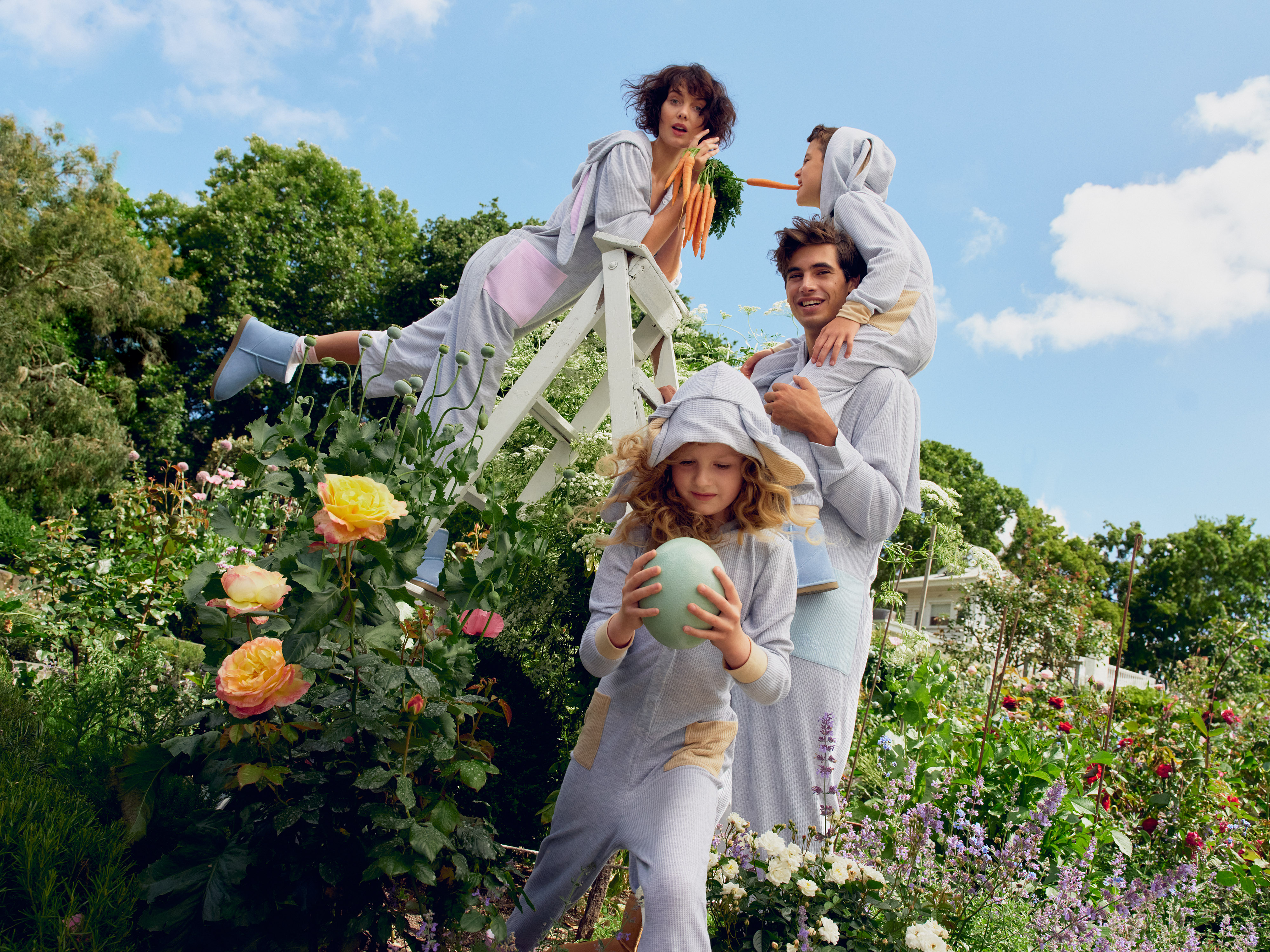 A family of four poses in a sunny garden wearing matching grey bunny-themed pajamas. The group is surrounded by roses and greenery.