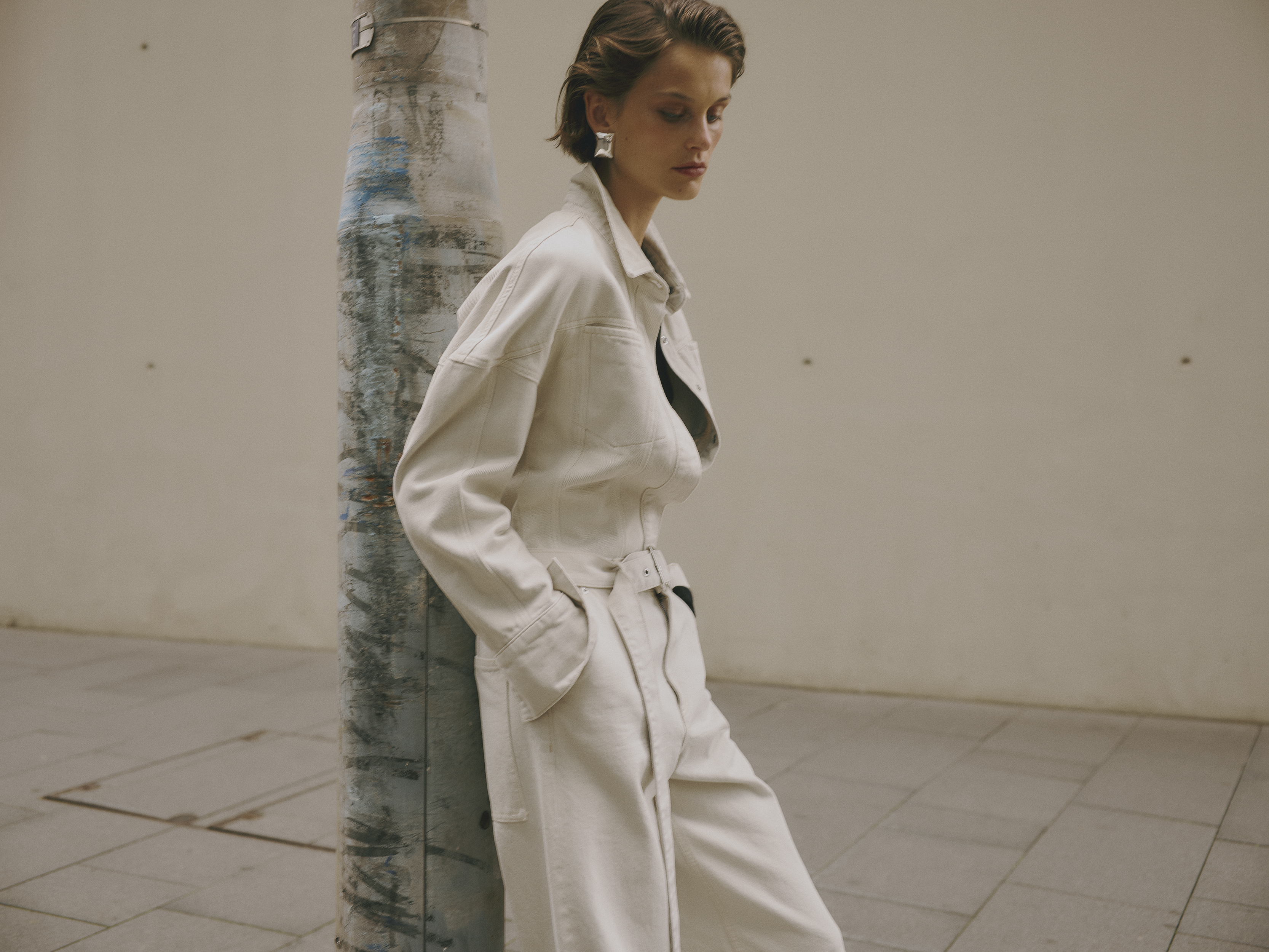 A slender woman with short hair poses in a beige jumpsuit, leaning against a pole on a city sidewalk in a fashion editorial.