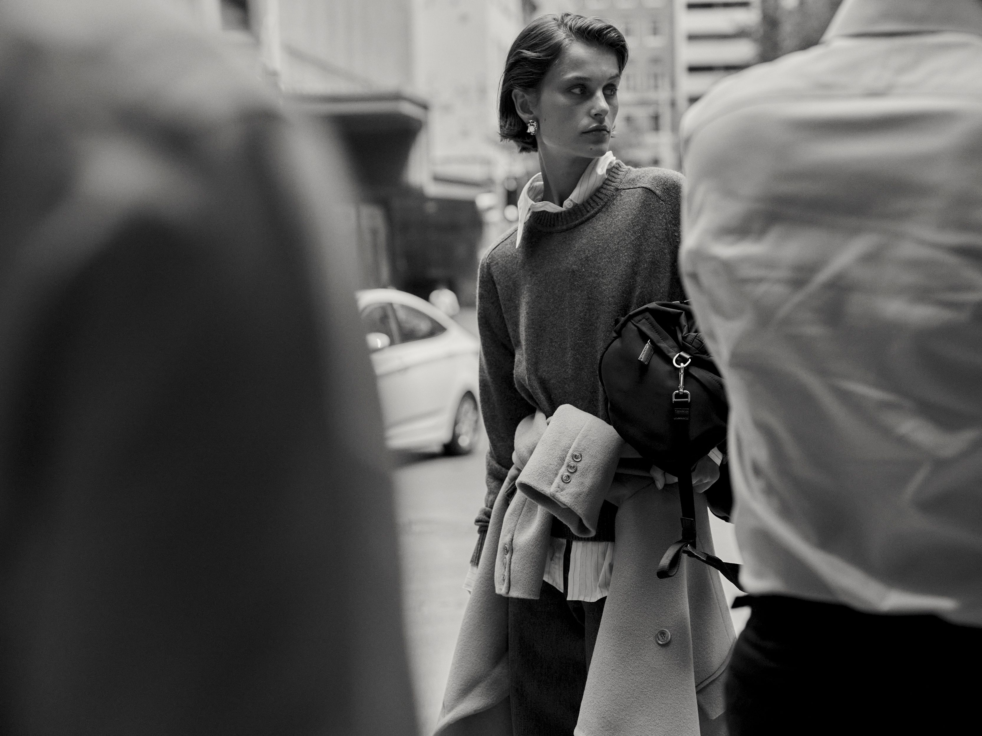Black and white fashion shot of a woman in a sweater and jacket walking in a city, framed by blurry pedestrians.