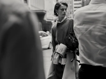 Monochrome street portrait of a woman in a knitted sweater holding a dark handbag with blurred figures framing the foreground.