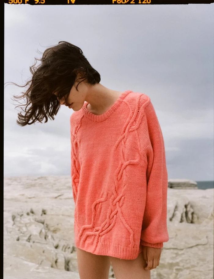 Model wearing a textured coral pink knit sweater outdoors with hair blowing in the wind against a pale rocky coastal landscape.