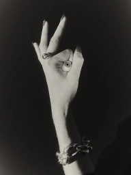 Close-up of a hand wearing a sculptural ring and chunky bracelet, holding a prosthetic eyeball, against a dark background.