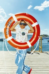 Young child holding a large orange lifebuoy around their head on a wooden jetty near sparkling blue water under a bright sky.