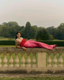 Woman in strapless red and pink gown reclining on a stone balustrade in a park by Adrian Price