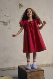 A young girl with curly red hair wearing a red dress standing on a wooden crate against a concrete wall