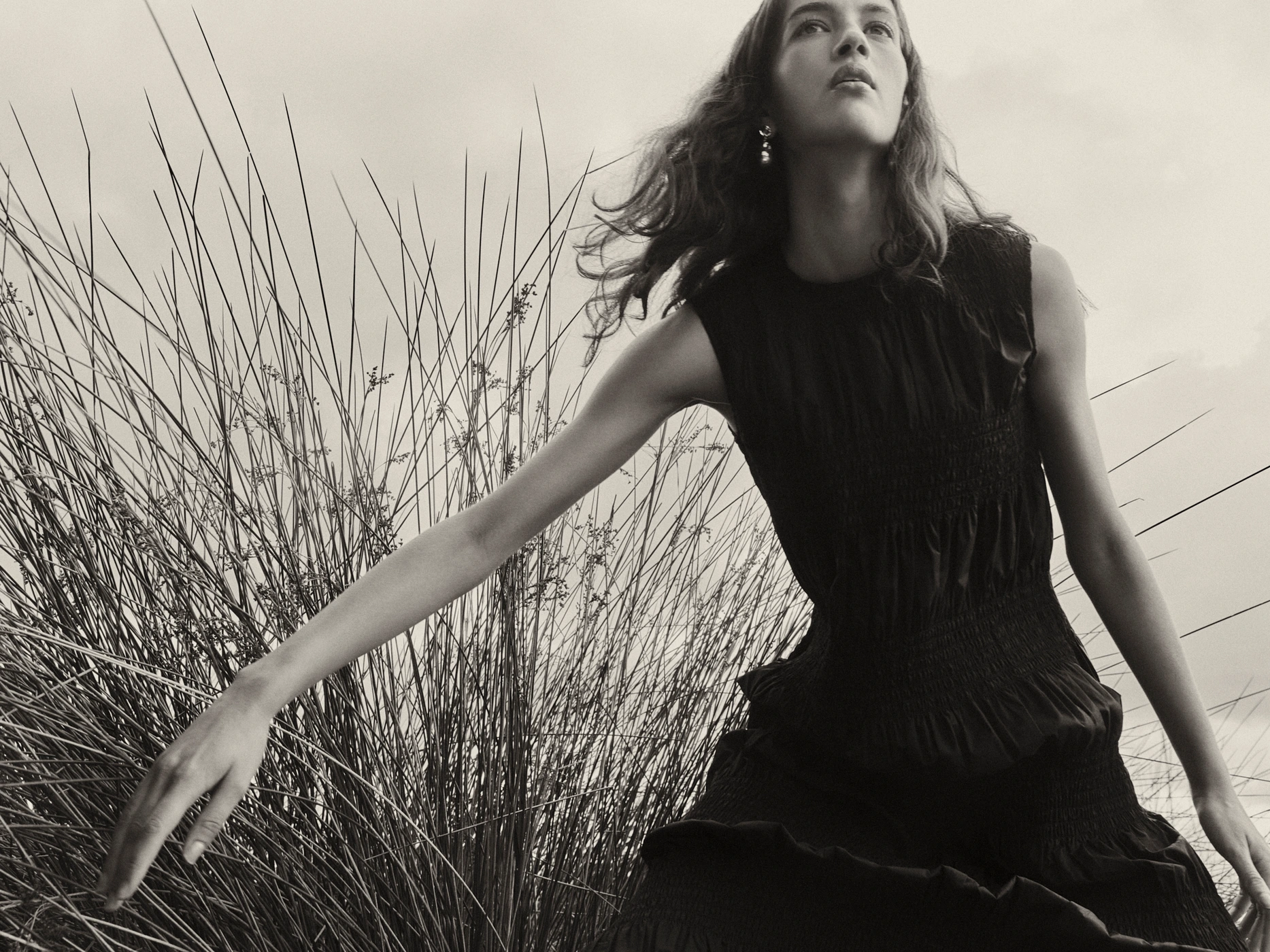 Model in a black sleeveless smocked dress posing among tall grass under an overcast sky by Adrian Price