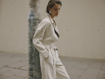 Woman wearing white belted jumpsuit leaning against a weathered concrete pillar in daylight