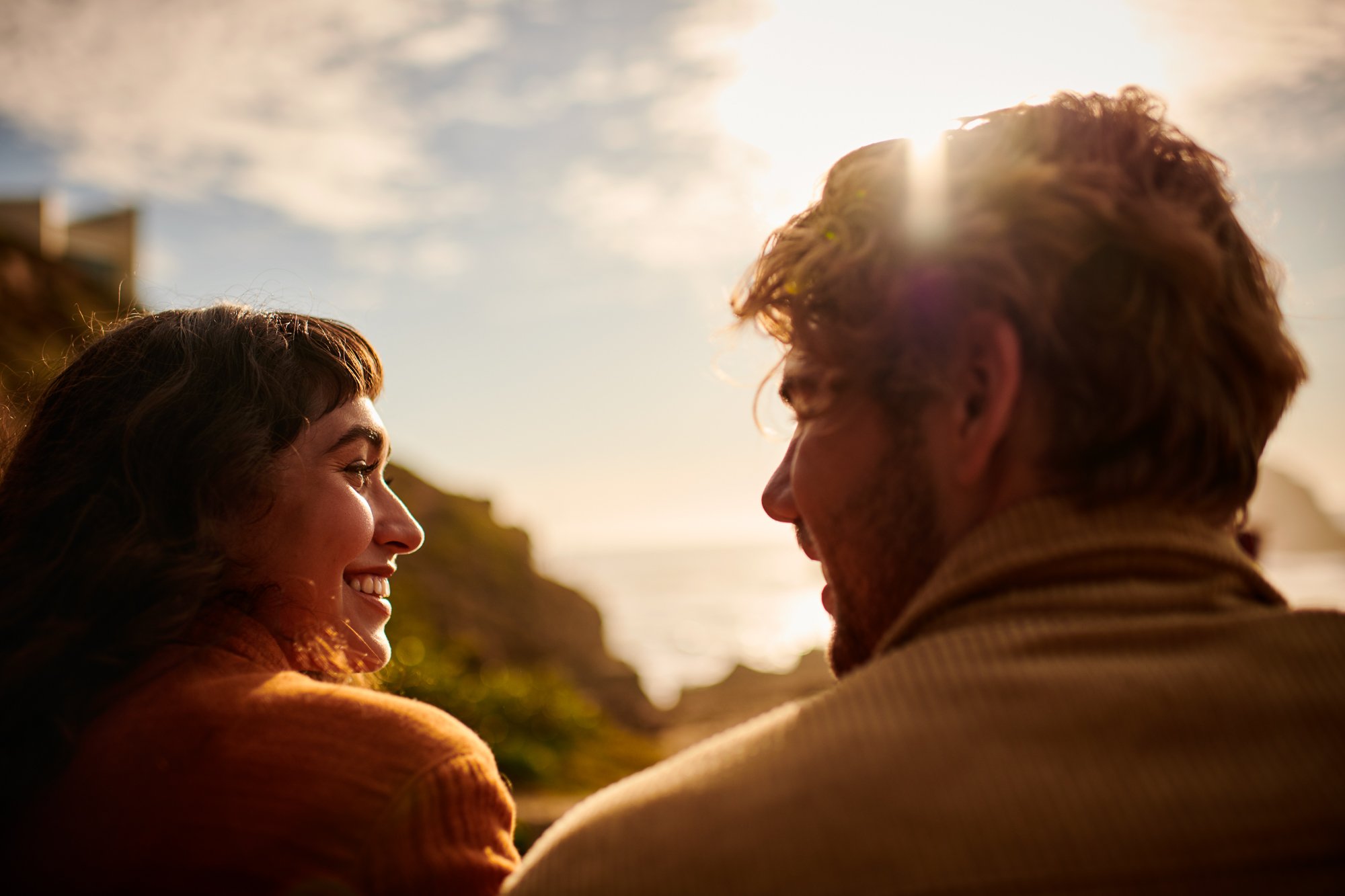 Woman and man smiling at each other with ocean background, bathed in warm sunlight creating a serene and romantic atmosphere.