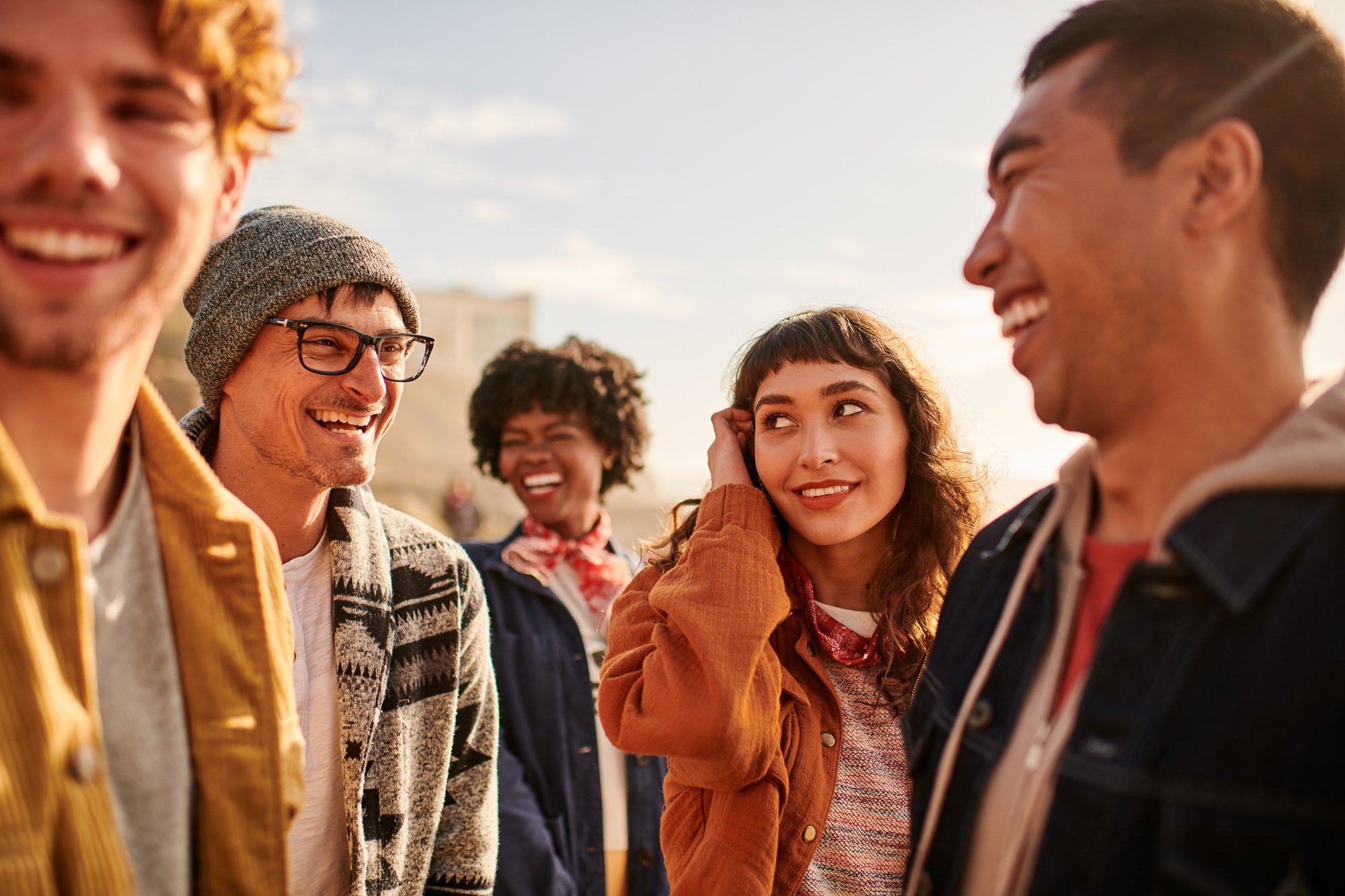 A group of five diverse young adults are outside, laughing in the sun. Close-up shot features casual clothing, with focus on joyful interaction.