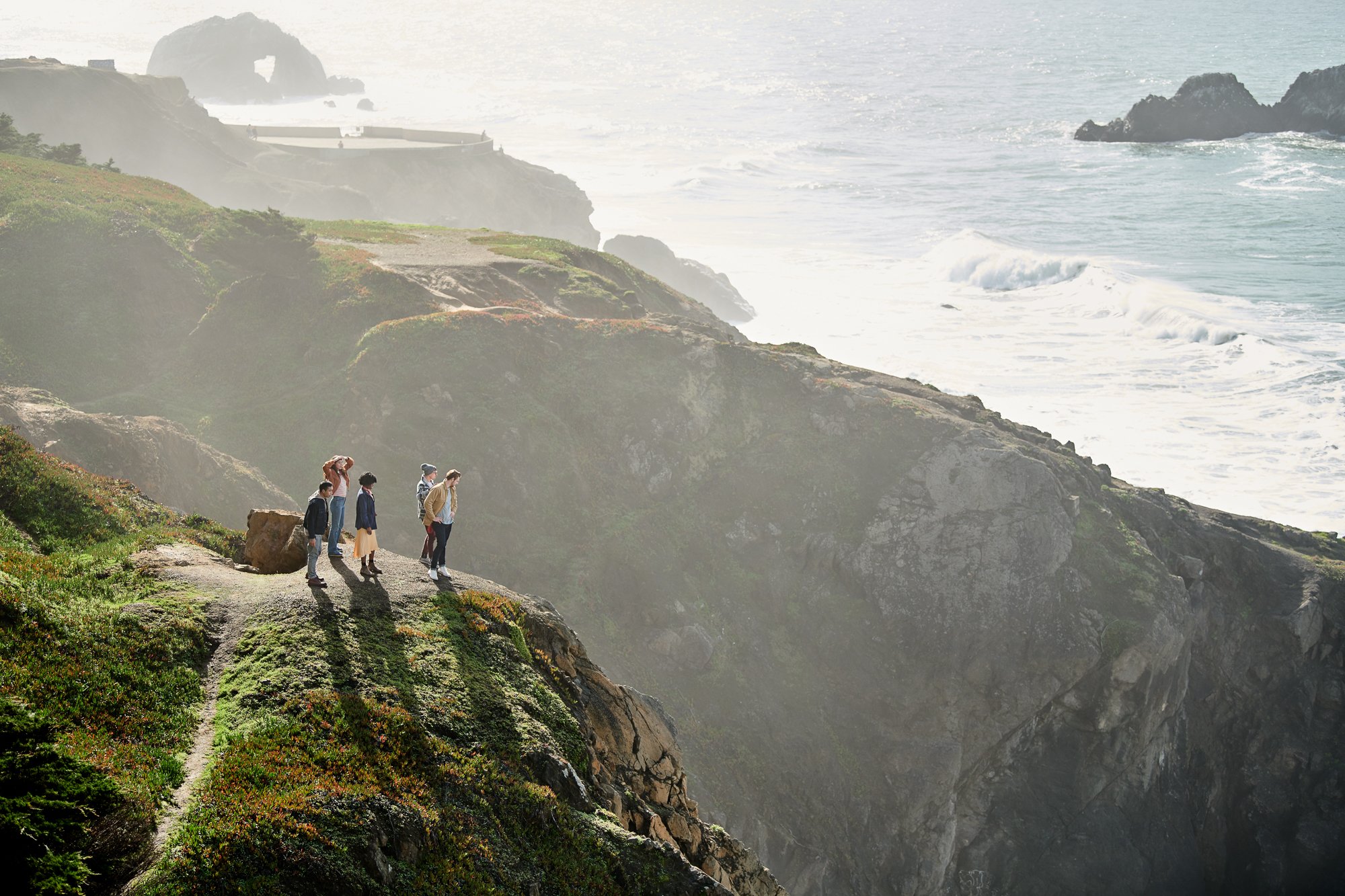 A group of young adults stand on a cliff overlooking the ocean on a sunny day, with rocky cliffs and crashing waves in the background.