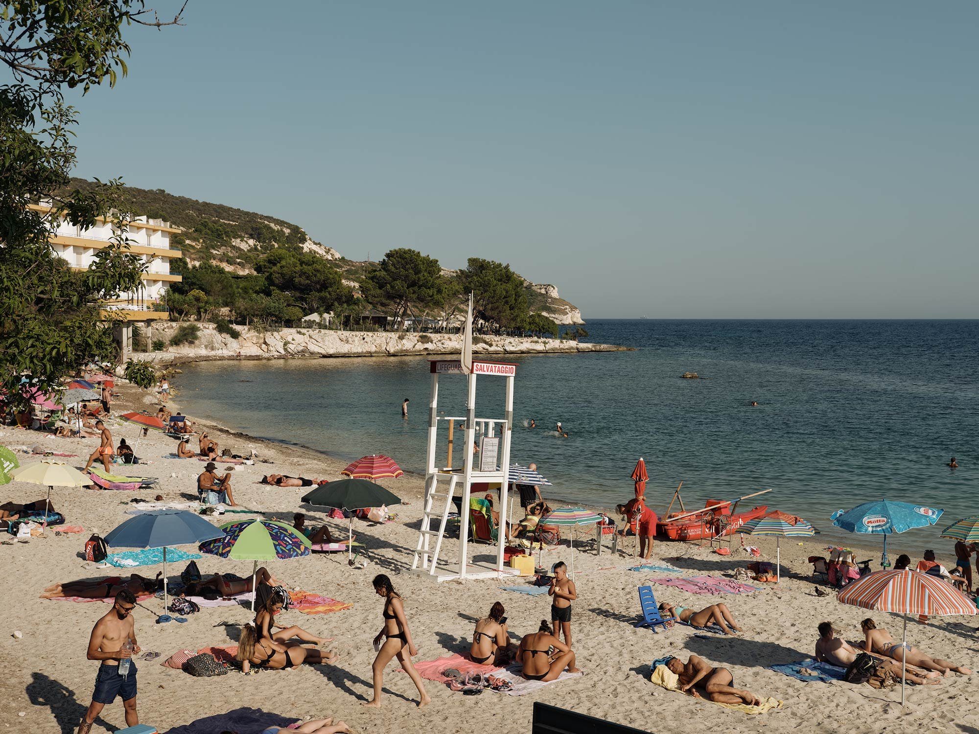 Sandy beach scene with people sunbathing, swimming, and relaxing near clear blue water, featuring lifeguard station and coastal vegetation.