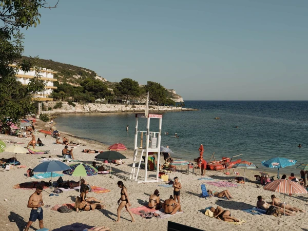 Sandy beach scene with people sunbathing, swimming, and relaxing near clear blue water, featuring lifeguard station and coastal vegetation.