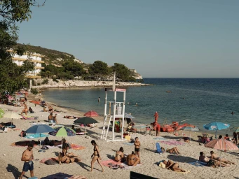 Wide shot of a crowded sandy beach with colourful umbrellas, a lifeguard tower, people sunbathing, and a coastal town in the background.