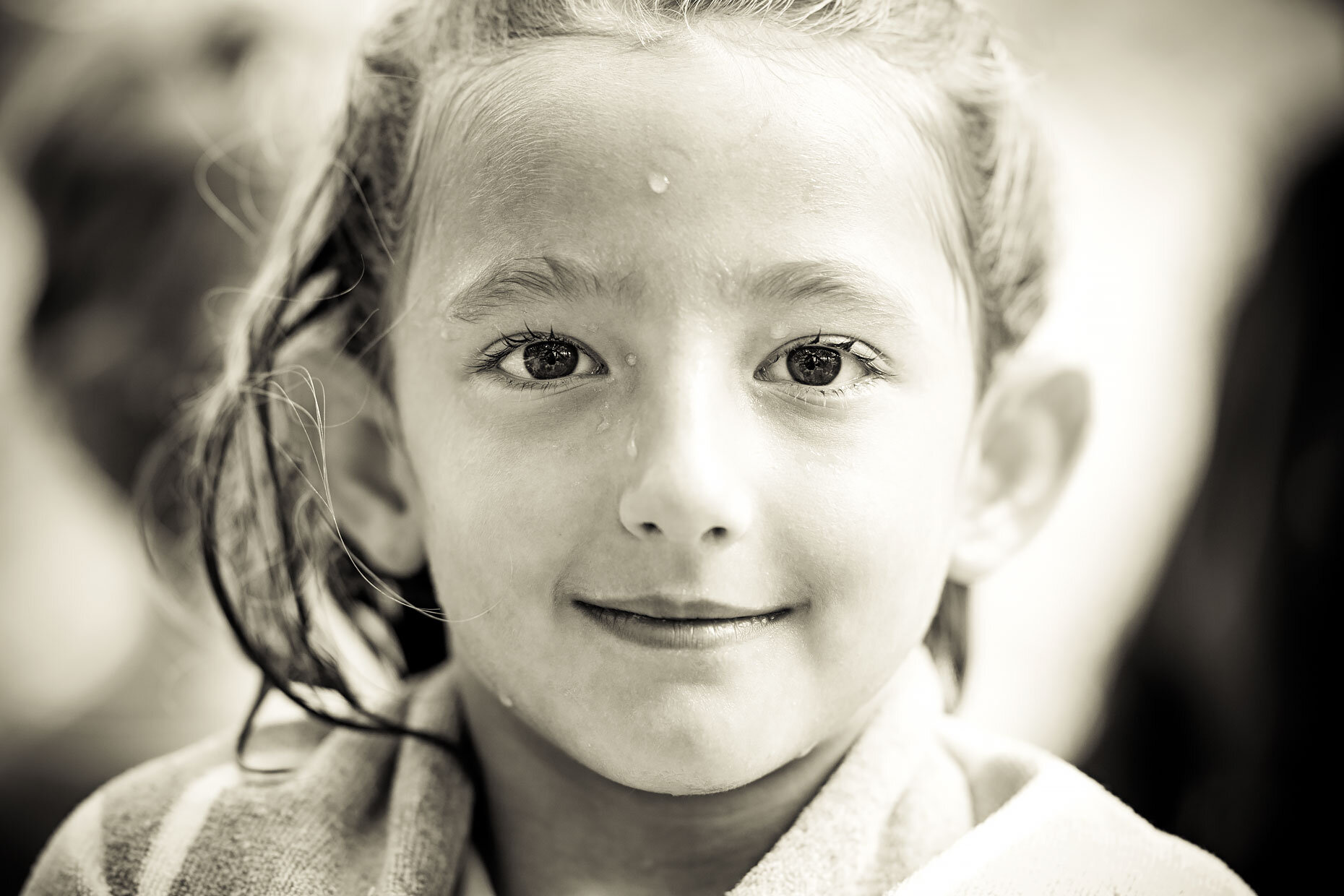 Close-up portrait of a young girl with wet hair and a striped towel, looking at the camera with a gentle smile in black and white.