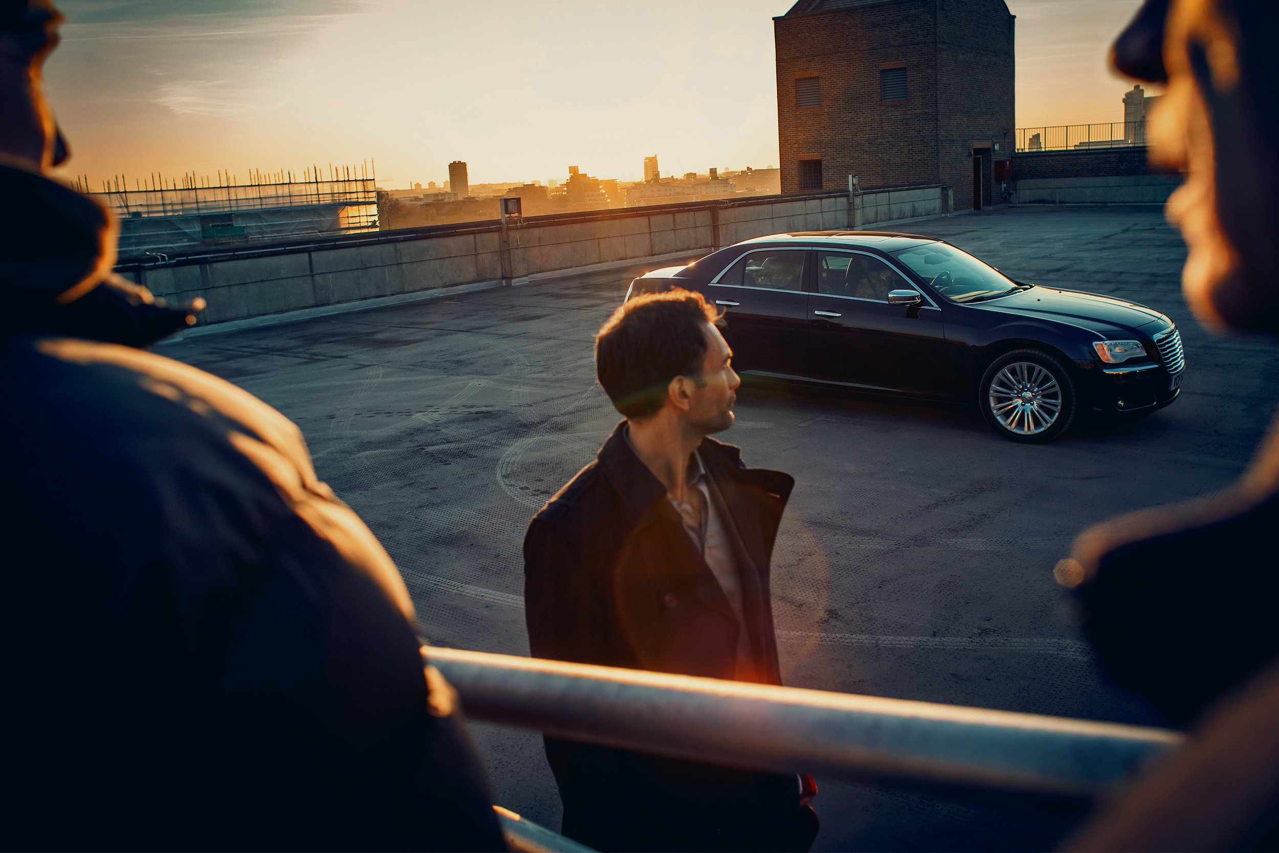 Adult man stands in urban setting with cityscape in the distance, a black car parked behind him, with two people in the foreground.