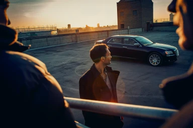 Man standing on a rooftop at sunset looking toward a parked dark sedan, framed by out-of-focus figures in the foreground