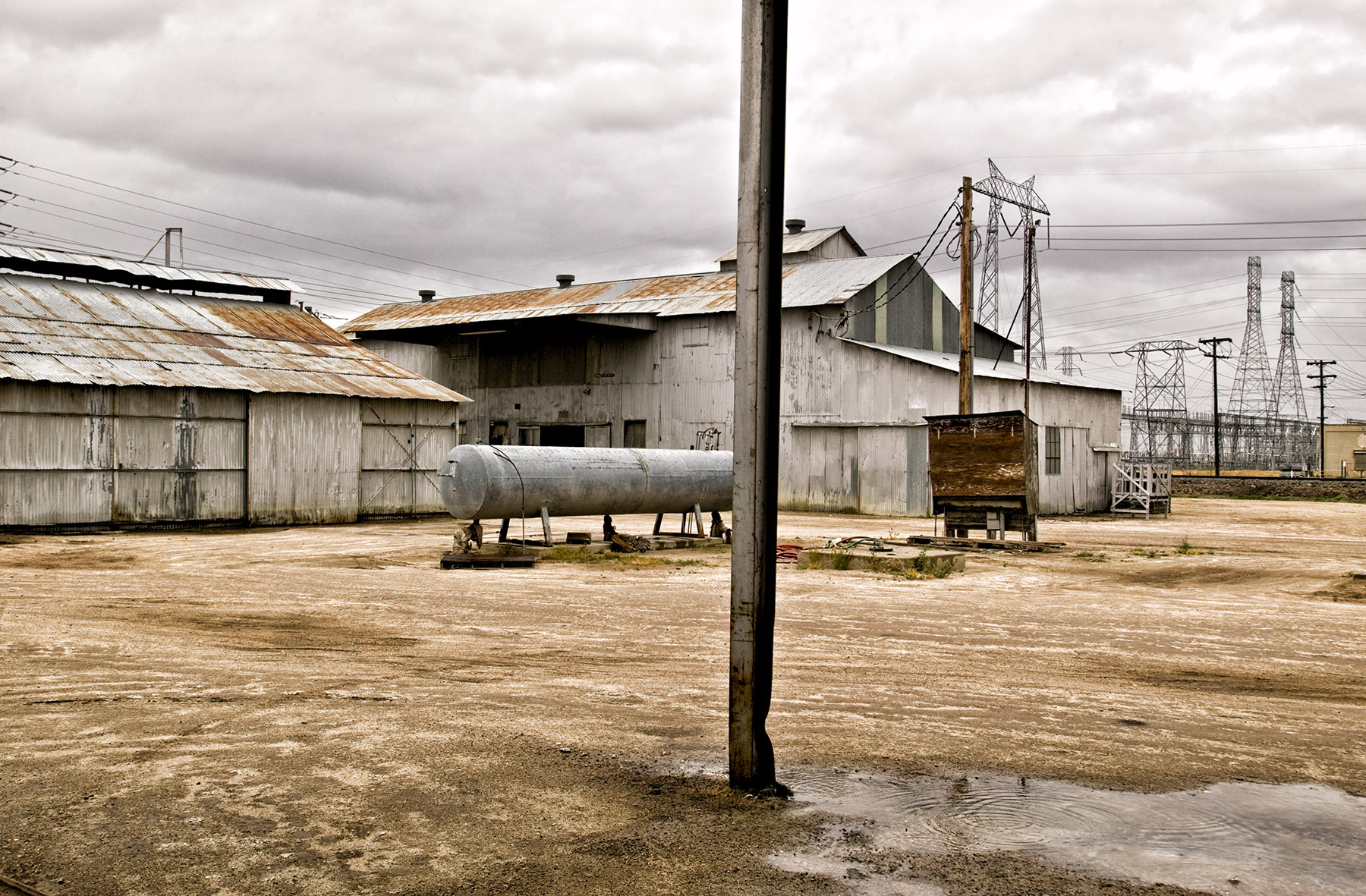 Industrial buildings under a cloudy sky, featuring corrugated metal structures, utility poles, and an aged, weathered aesthetic.