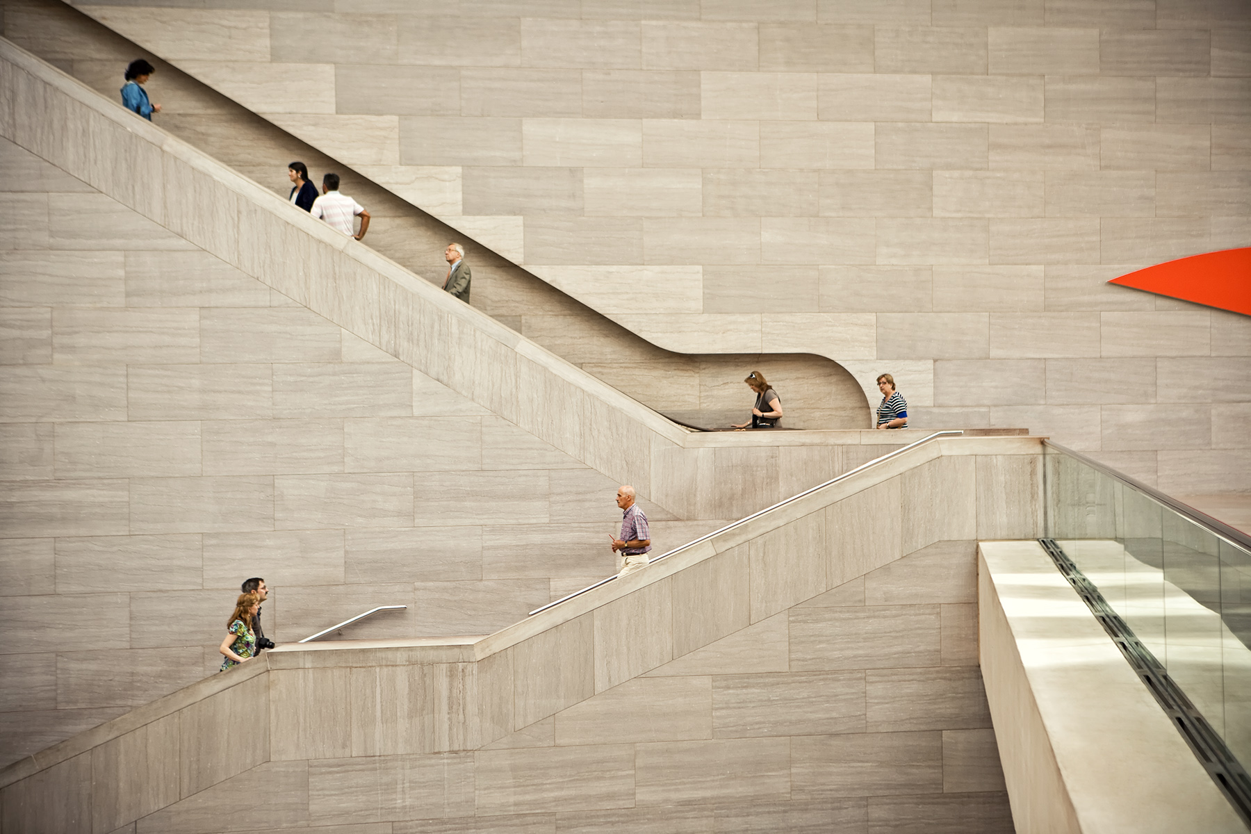 Wide shot captures multiple people traversing a multi-level staircase with a light stone wall background and orange architectural accent.