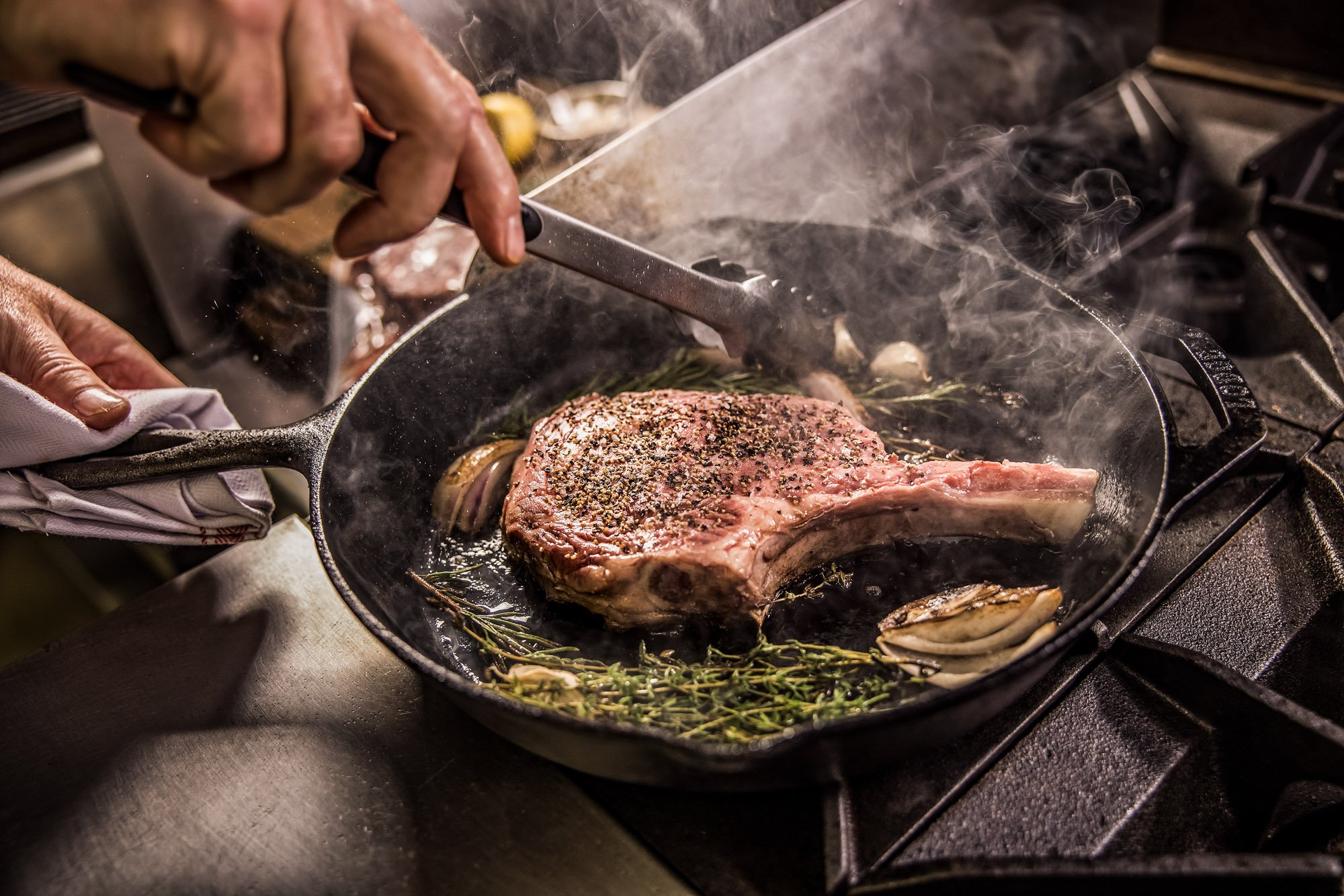 A chef prepares a bone-in steak in a cast iron skillet with tongs, aromatics, and steam in a commercial kitchen setting.