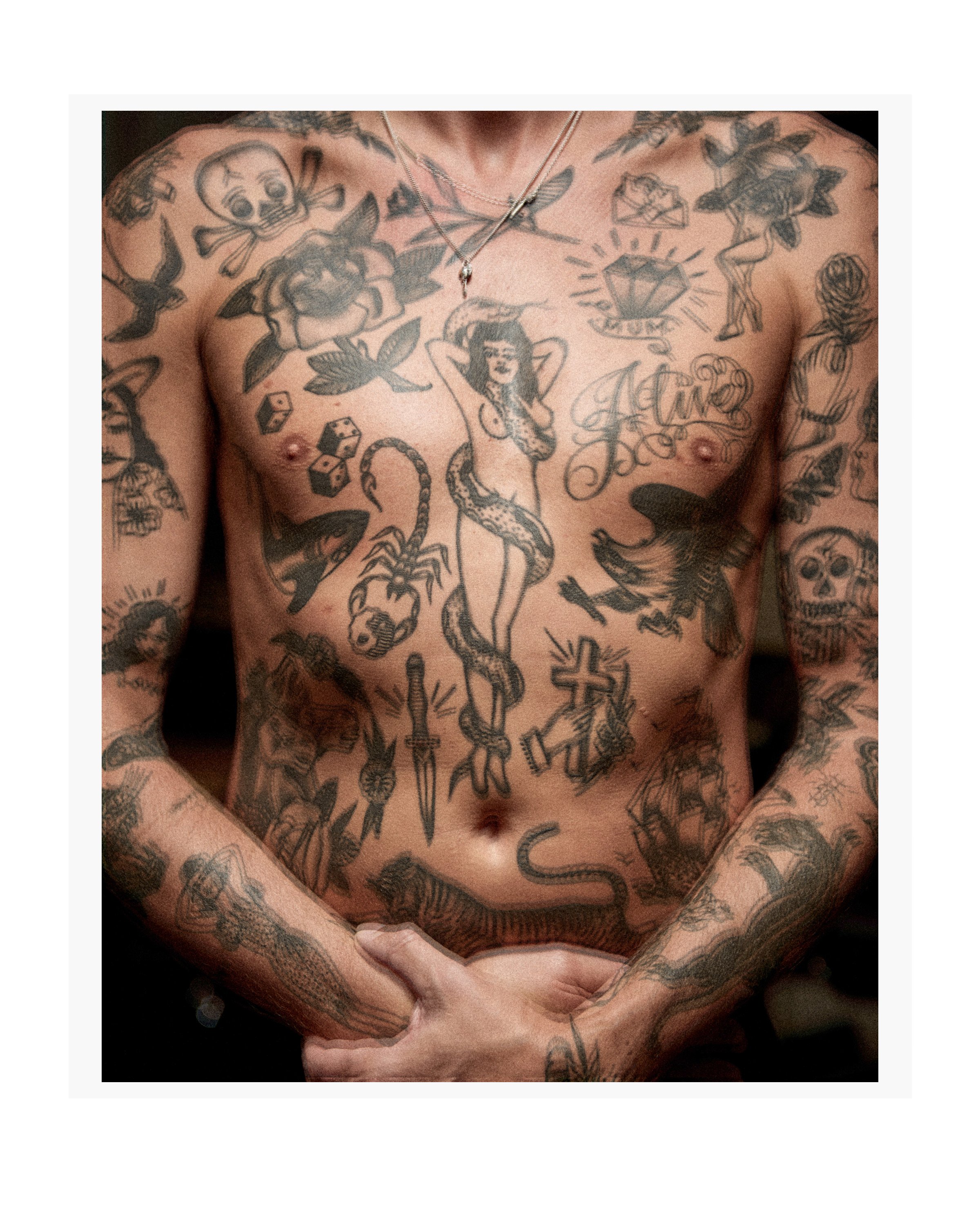 Torso of fair-skinned adult male covered with dense black ink tattoos of skulls, flowers, animals, and a nude woman with a snake.