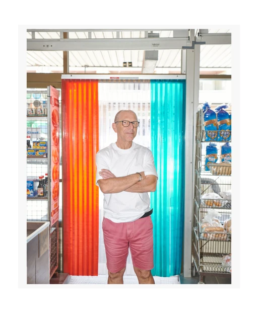 Elderly man in glasses and shorts stands with arms crossed in a shop next to orange and teal plastic strip curtains and retail shelves.
