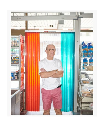 Elderly man in glasses and shorts stands with arms crossed in a shop next to orange and teal plastic strip curtains and retail shelves.