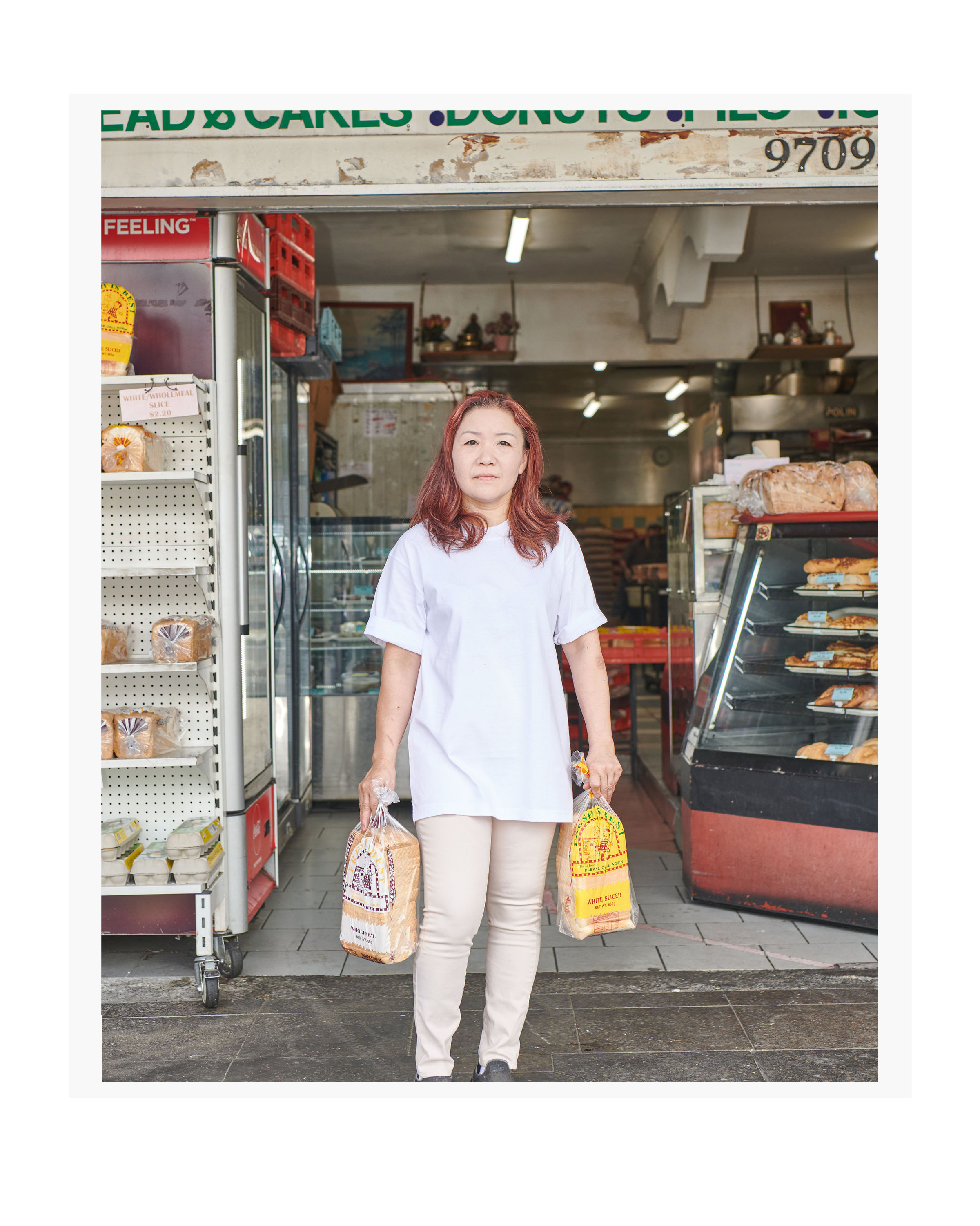 Woman in front of bakery holding two loaves of bread in plastic bags, wearing a white t-shirt and beige pants, standing outdoors.