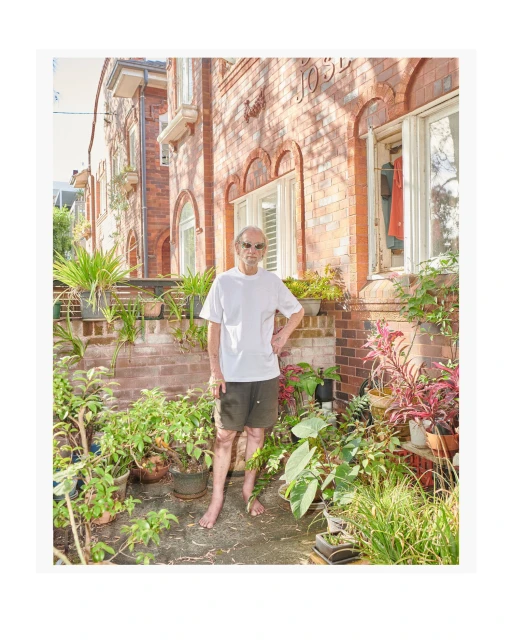 A man stands barefoot among potted plants in front of a brick building with windows. He wears a white t-shirt and dark shorts.