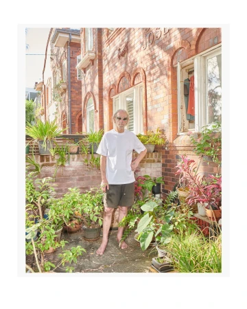 A man stands barefoot among potted plants in front of a brick building with windows. He wears a white t-shirt and dark shorts.