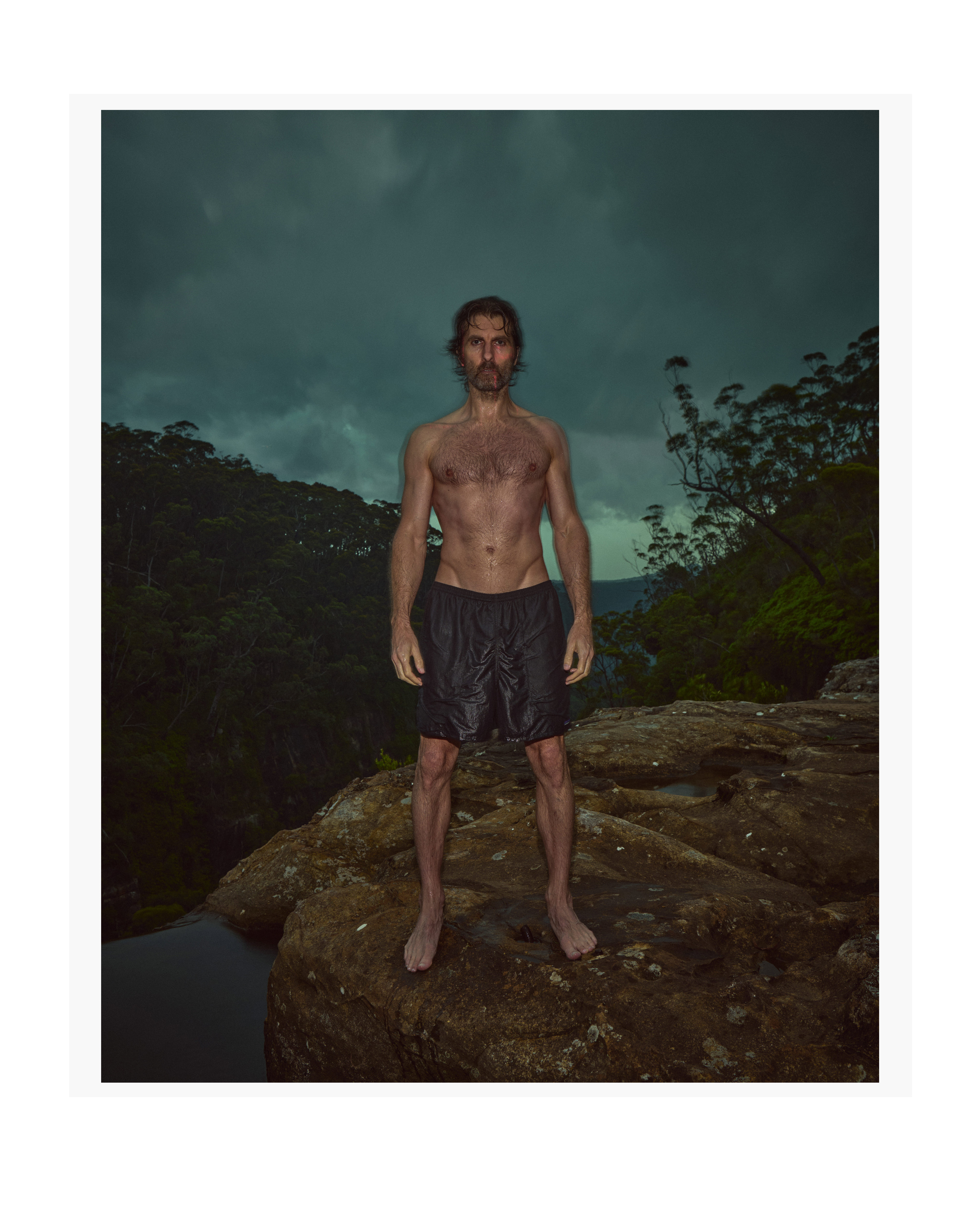 Adult male stands on a rocky outcrop overlooking a forested valley under an overcast sky, wearing black shorts, captured in portrait orientation.