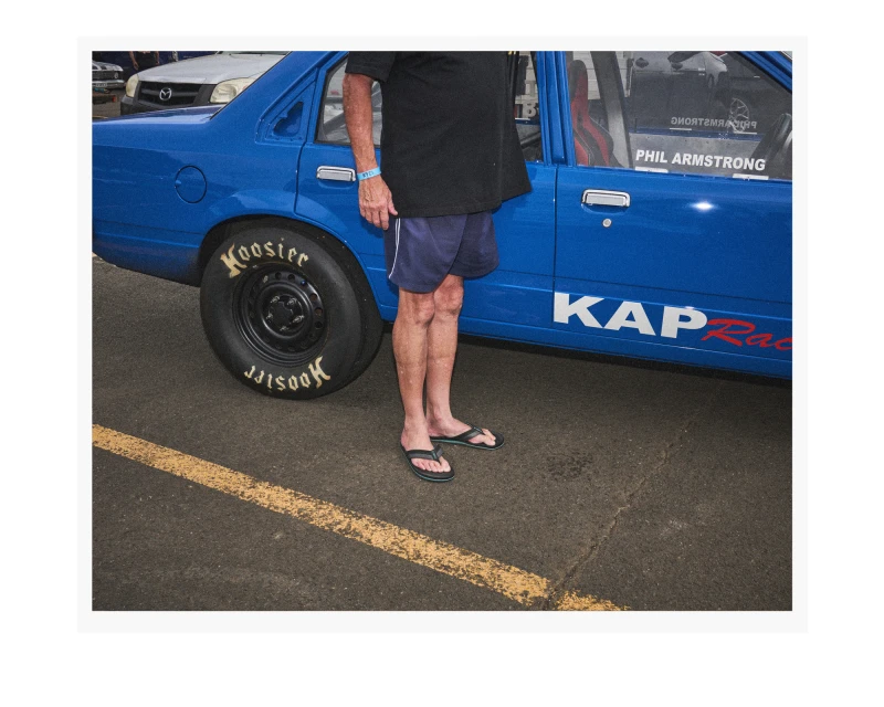 Blue racing car with a black tire and an adult in navy shorts and flip-flops standing near it, outdoors on an asphalt surface.